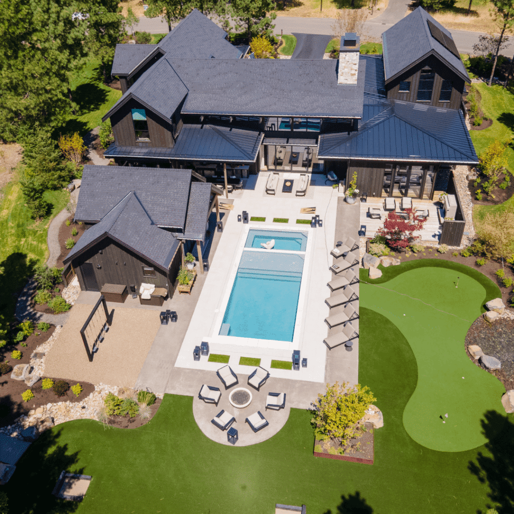 Aerial view of a modern home with a central swimming pool, lounge chairs, outdoor seating areas, a fire pit, and a putting green surrounded by landscaped gardens and trees.