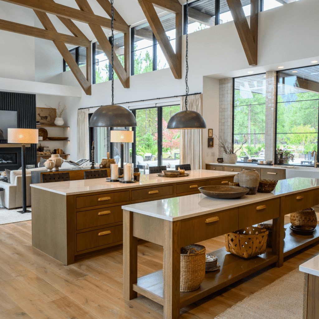 A spacious, modern kitchen with wood beams, large windows, and two islands with white countertops. Pendant lights hang above, and baskets and decor accentuate the warm, open space with greenery visible outside.