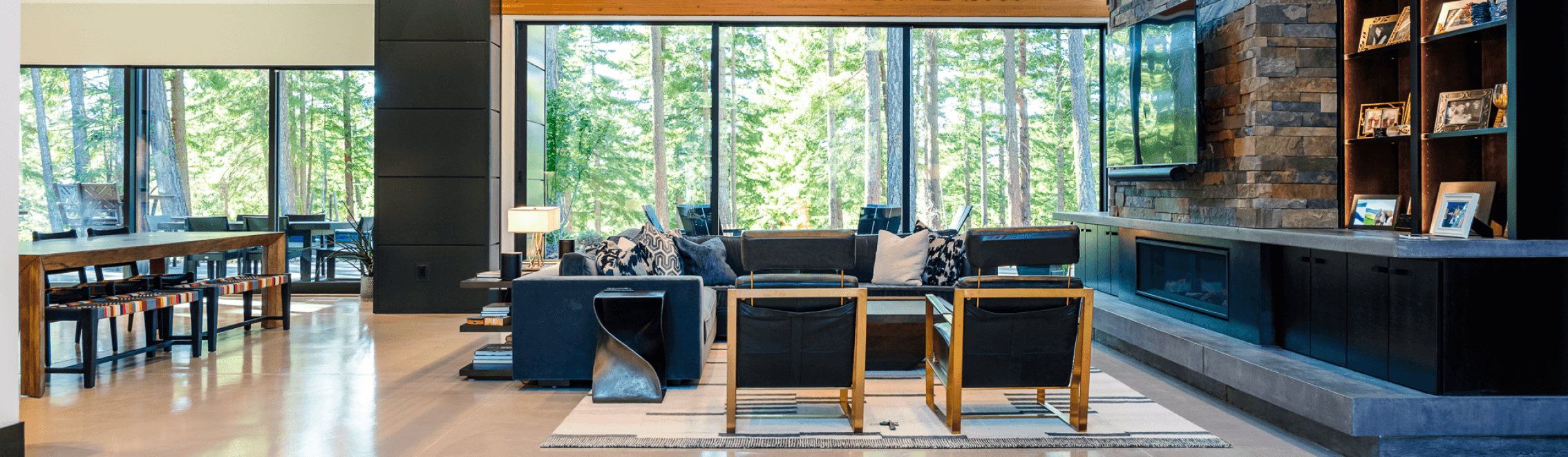 Modern living room with floor-to-ceiling windows, forest view, contemporary furniture, a long dining table, built-in fireplace with stone surround, and family photos on display. Natural light fills the spacious, open layout.