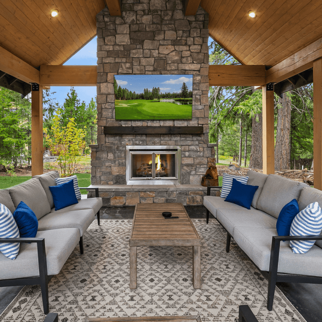 Covered outdoor patio with a stone fireplace, TV above the mantle, two gray sofas with blue and striped pillows, a wooden coffee table, and a patterned rug, surrounded by trees and greenery.