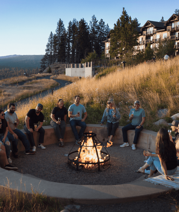 A group of people sit around a fire pit outdoors, surrounded by grass and trees, with drinks in hand. Buildings and a hillside are visible in the background under a clear sky.