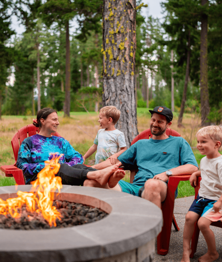 A family of four sits in red chairs around a fire pit outdoors, smiling and laughing together. Tall trees and greenery are visible in the background.