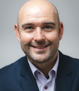 A smiling, bald man with a trimmed beard, wearing a dark blazer over a light shirt, poses in front of a plain, light background.