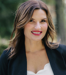 A woman with shoulder-length brown hair, wearing a black blazer and white top, smiles outdoors with a blurred green background.