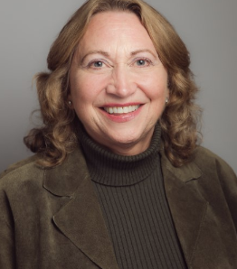Smiling woman with wavy light brown hair, wearing a green turtleneck sweater and an olive green suede jacket, posed against a plain gray background.