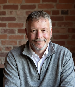 A middle-aged man with short gray hair and a beard, wearing a light gray zip-up sweater over a white shirt, smiles while sitting in front of a brick wall.