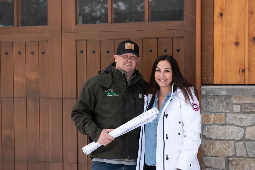 A man wearing a dark jacket and baseball cap holds rolled-up blueprints, standing next to a smiling woman in a white coat. They are posing in front of a wooden garage door with stone accents.