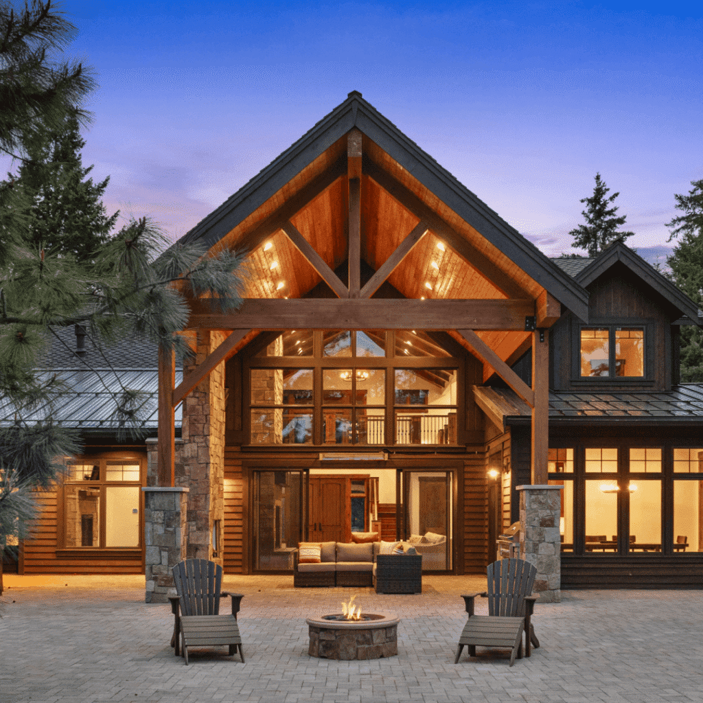 A large, modern cabin-style house with tall wooden beams, expansive windows, and warm glowing lights at dusk. Two Adirondack chairs surround a fire pit on a stone patio in the foreground.
