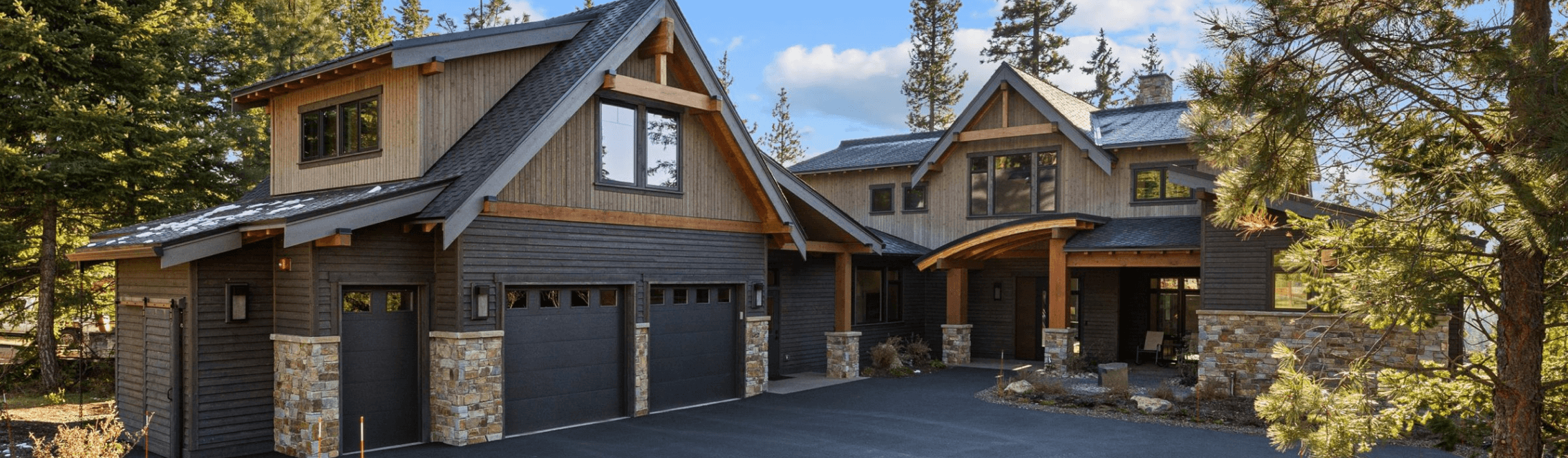 A modern two-story house with dark wood siding, stone accents, and a double garage, surrounded by tall pine trees under a clear blue sky.
