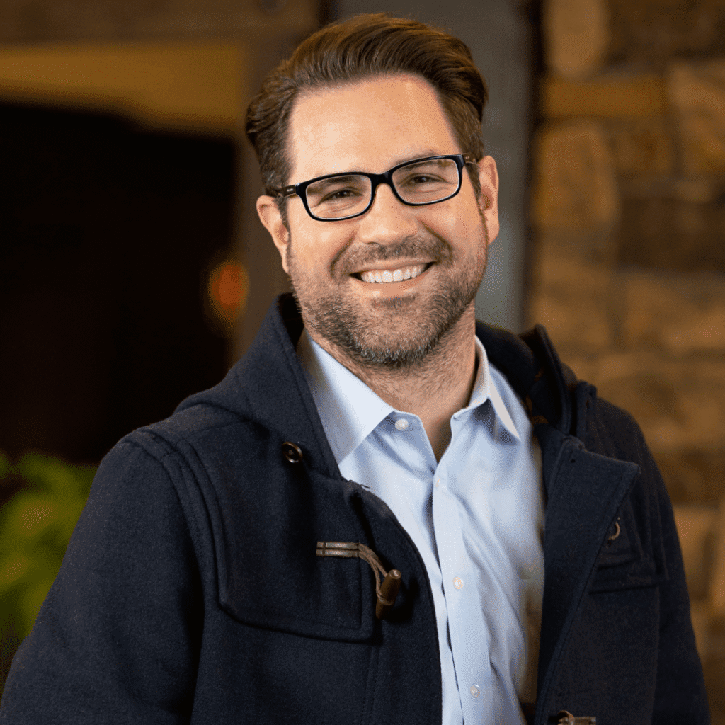 A man with dark hair and glasses smiles at the camera. He is wearing a navy jacket over a light blue collared shirt, standing indoors with a stone wall and soft lighting in the background.