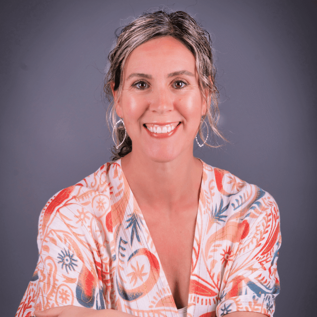 A woman with wavy, shoulder-length hair and hoop earrings smiles at the camera. She is wearing a patterned, colorful blouse and is posed in front of a plain gray background.
