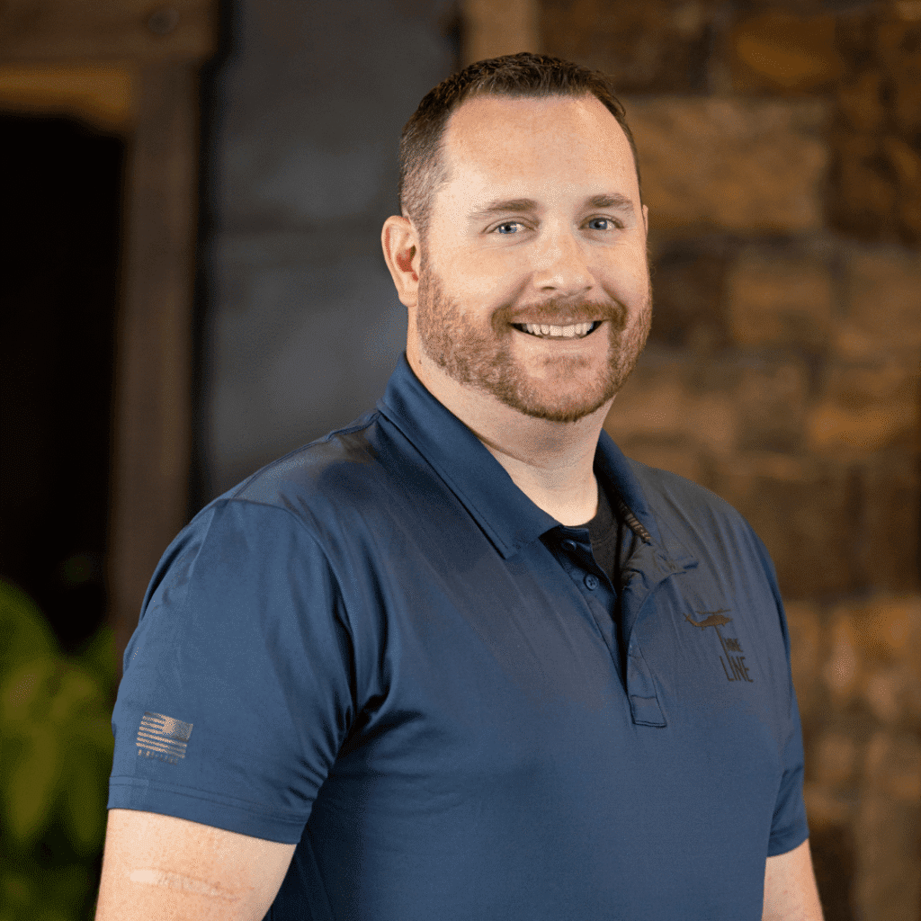 A man with short brown hair and a beard, wearing a blue polo shirt, smiles while standing indoors in front of a stone wall.
