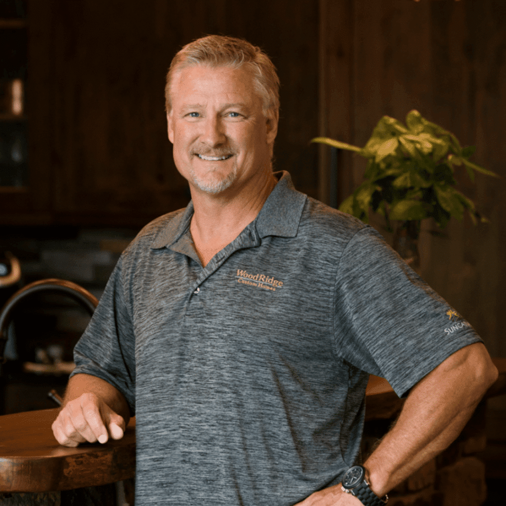 A middle-aged man with short gray hair and a beard smiles while leaning on a wooden counter. He wears a gray polo shirt with embroidered logos and stands in a warmly lit room with wood accents and a plant in the background.