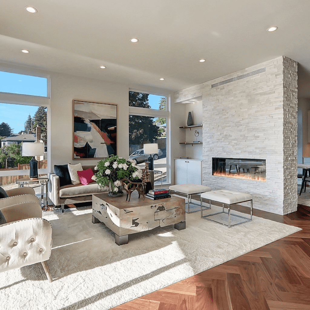Modern living room with large windows, a white stone fireplace, abstract art, neutral furniture, a wooden coffee table with flowers, and natural light filling the space.
