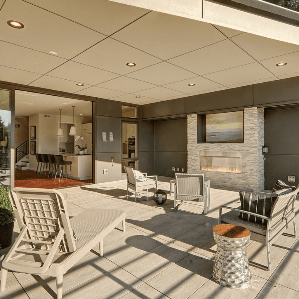 Modern outdoor patio with light wooden flooring, lounge chairs, and a seating area facing a stone fireplace with a landscape painting above it. The space opens to an indoor kitchen and dining area through glass doors.