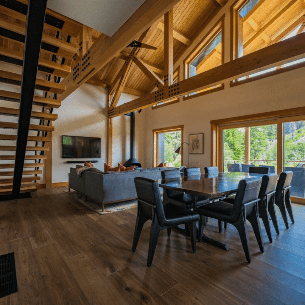 Modern open-concept living and dining area with exposed wooden beams, large windows, a wooden staircase, gray sofa with orange pillows, black dining chairs, and views of greenery outside.