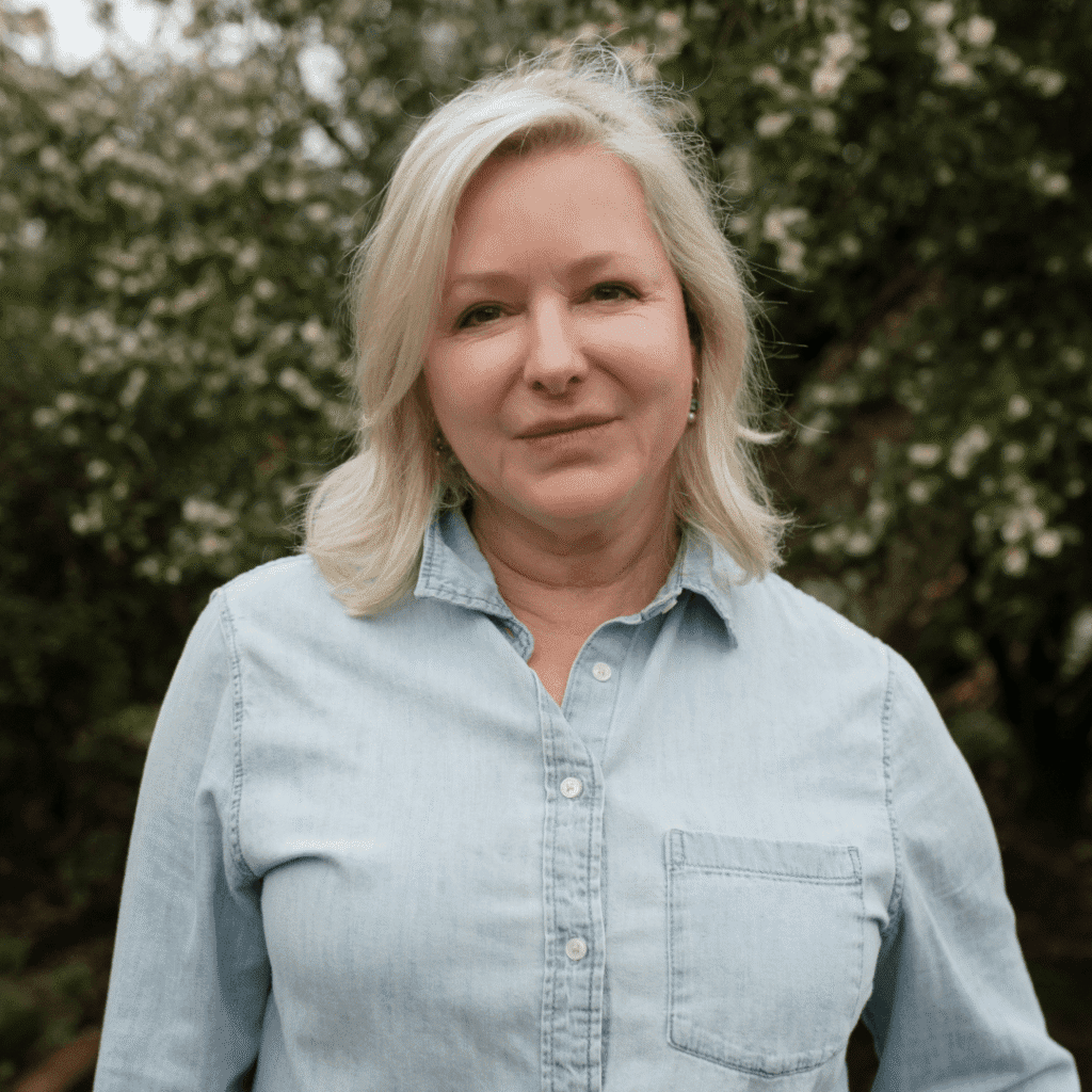 A woman with shoulder-length blonde hair wearing a light blue denim shirt stands outdoors in front of green foliage and flowering plants, looking calmly at the camera.
