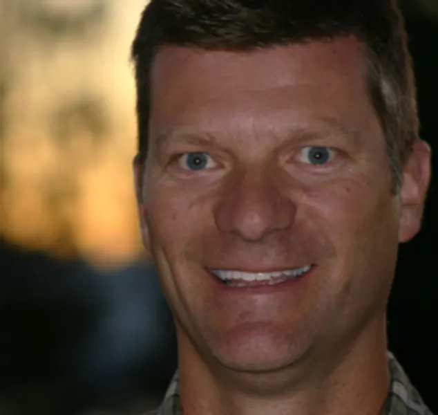 A man with short brown hair smiles at the camera; he is outdoors with a blurred sunset and trees in the background.