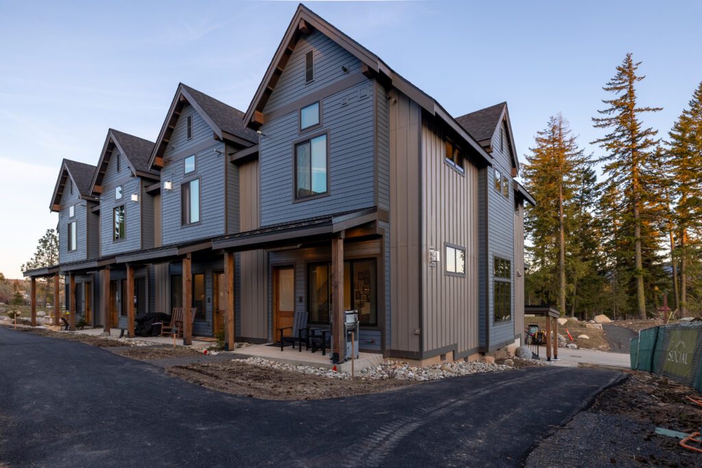 Row of modern, two-story townhouses with gray siding and large windows, set along a paved road with some construction materials and a few trees in the background.