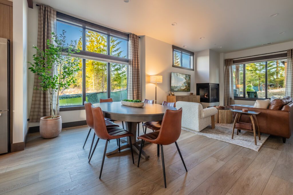 Modern, cozy living and dining area with large windows, wooden floor, round dining table with brown chairs, sofa, armchair, small fireplace, indoor plant, and views of trees outside. Natural light fills the space.
