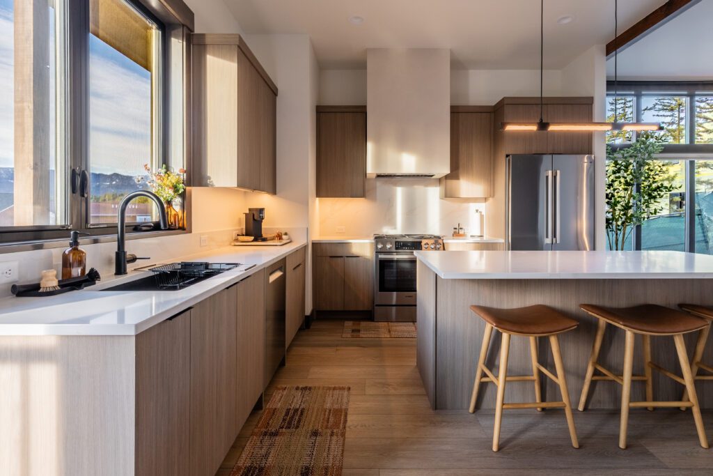 Modern kitchen with light wood cabinets, white countertops, a large window, stainless steel appliances, an island with two wooden stools, and warm natural light illuminating the space.
