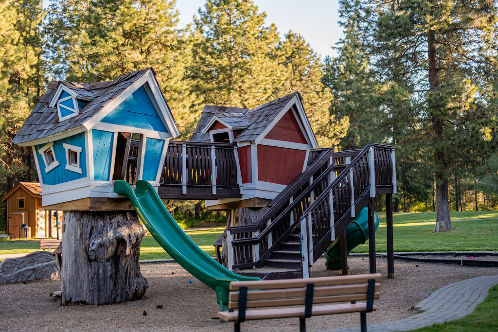 A whimsical playground structure with two colorful crooked playhouses, green slides, and stairs, set in a park with trees and grass. A bench sits in the foreground.