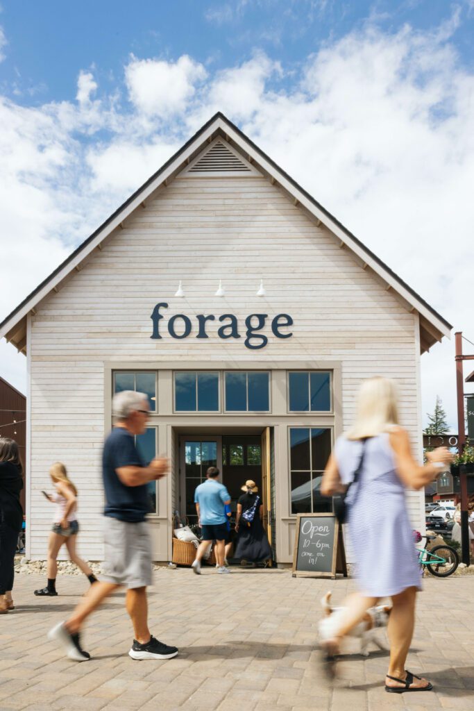People walk in front of a light-colored building with a gabled roof and large windows. The building has the word forage above the entrance and a sign outside that reads Open 11–6pm. The sky is partly cloudy.
