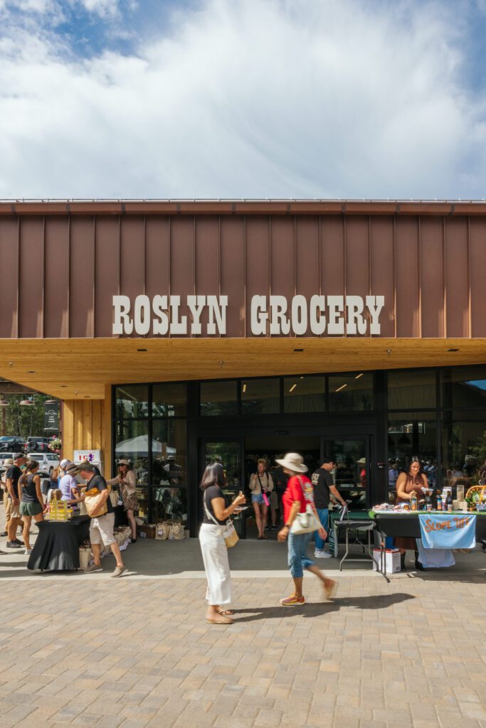 People gather outside Roslyn Grocery, a modern building with a wooden overhang. Several tables are set up with goods and displays, and the sky is partly cloudy.