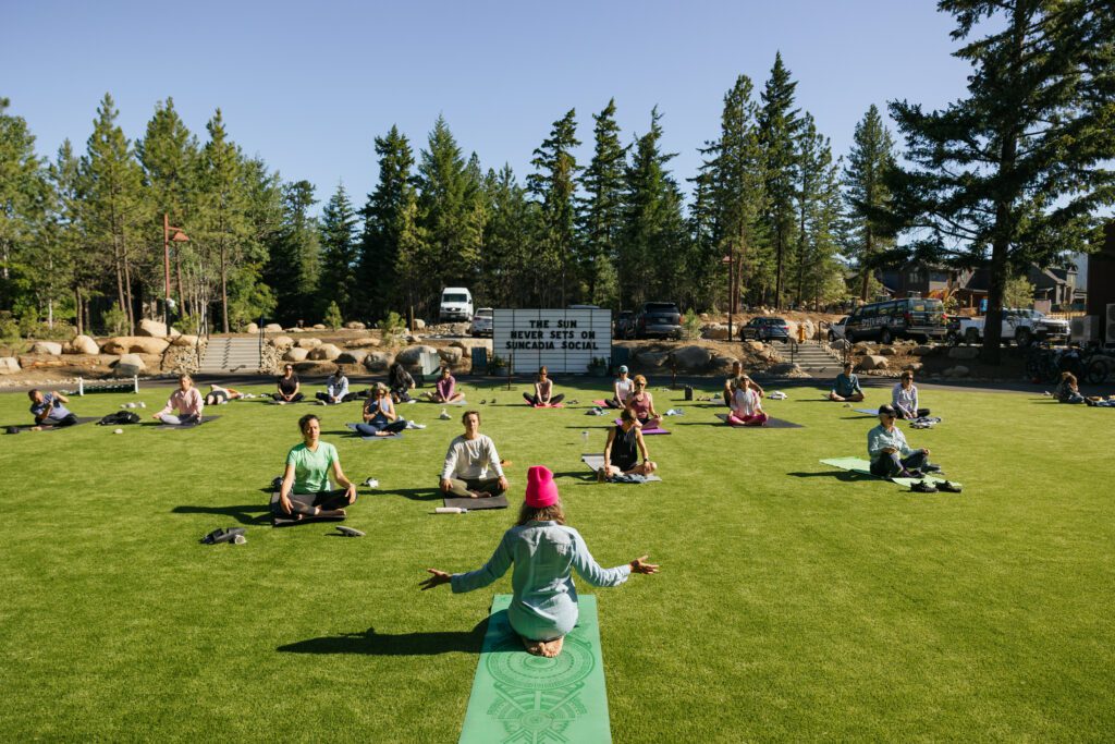 A group of people practices yoga outdoors on a grassy lawn, sitting on mats in a circle with an instructor at the front. Tall trees surround the area, and a sign is visible in the background.