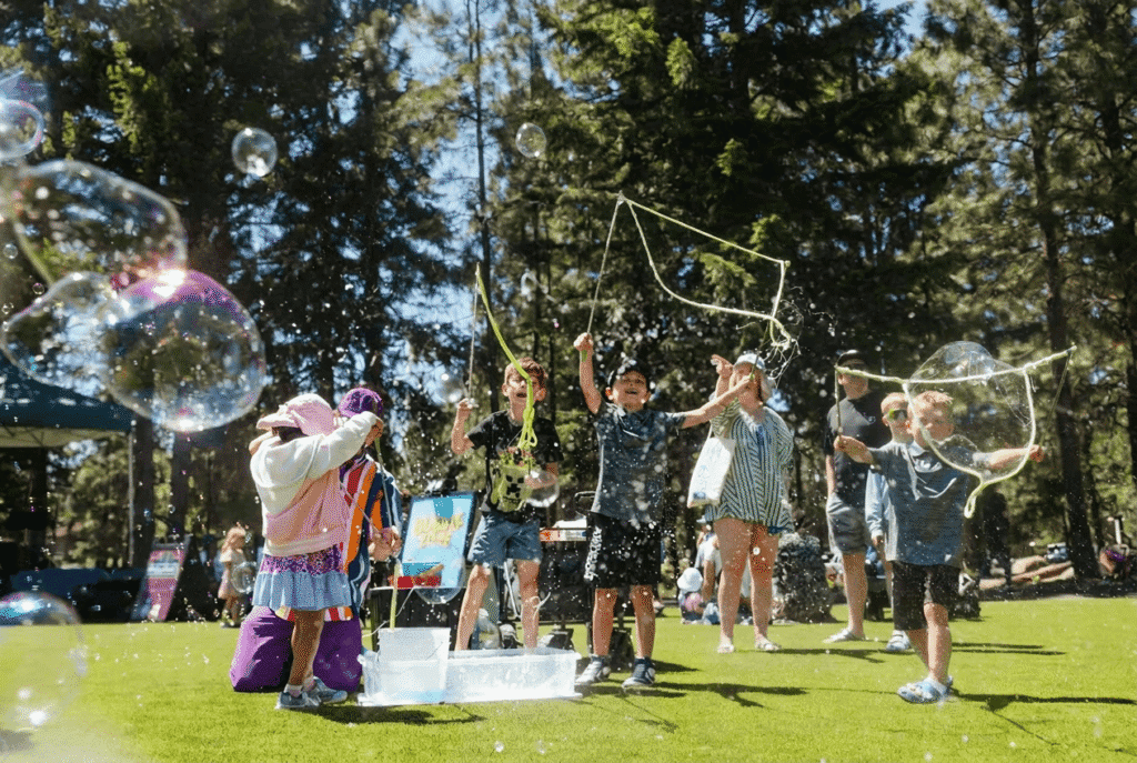 A group of children and adults play with giant bubbles on a grassy field surrounded by tall trees, enjoying a sunny day outdoors. Some kids wave bubble wands while others watch bubbles float in the air.