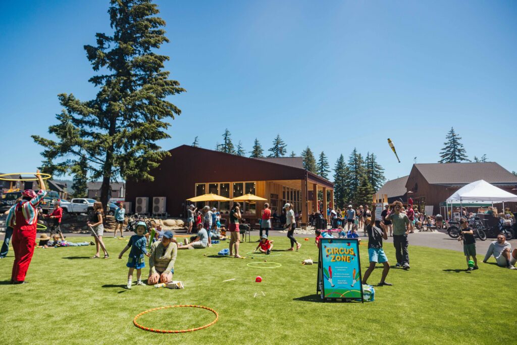 People of all ages enjoy outdoor activities on a sunny day at Spring events at Suncadia, with hula hoops, a “Circus Zone” sign, and surrounding buildings. Children play as adults supervise and socialize in the lively grassy area.