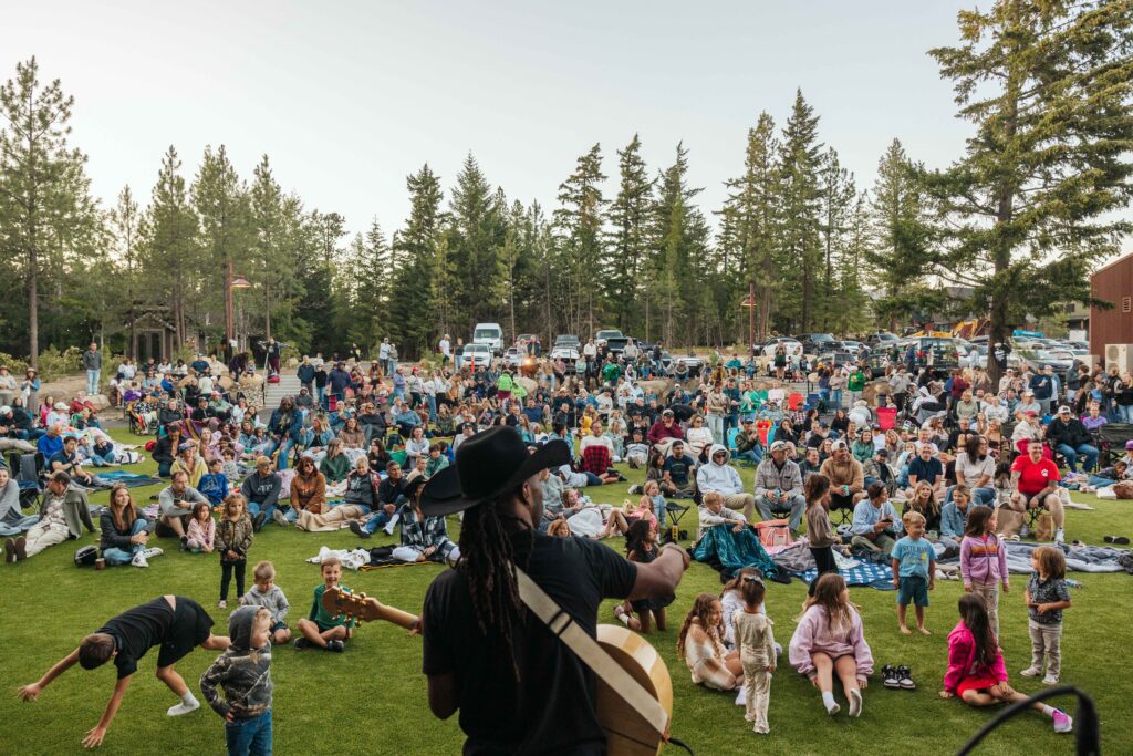 A musician plays guitar on an outdoor stage at Spring events at Suncadia, facing a large crowd sitting on blankets and chairs spread across a grassy field, with children in front and tall trees in the background.