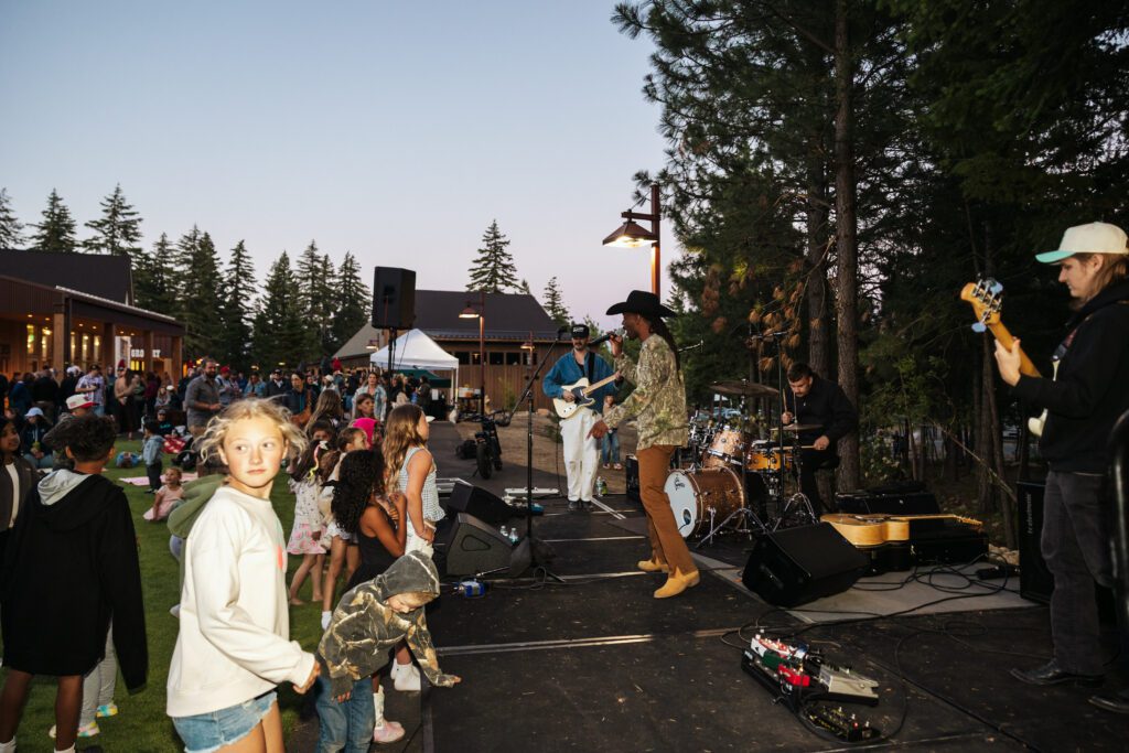 A band performs on an outdoor stage near a building at dusk while children and adults gather and dance on the grass. Tall pine trees and string lights create a festive atmosphere.