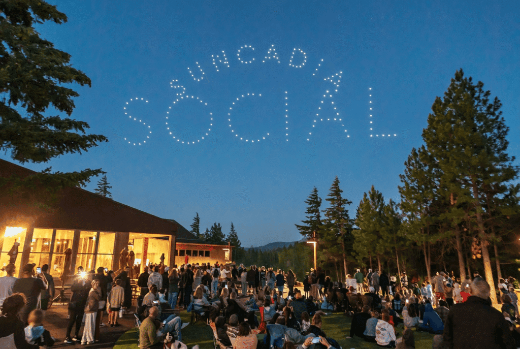 A large crowd gathers on a grassy area at dusk outside a building, watching drones form the words SUNCADIA SOCIAL in the clear blue sky above a forested landscape.