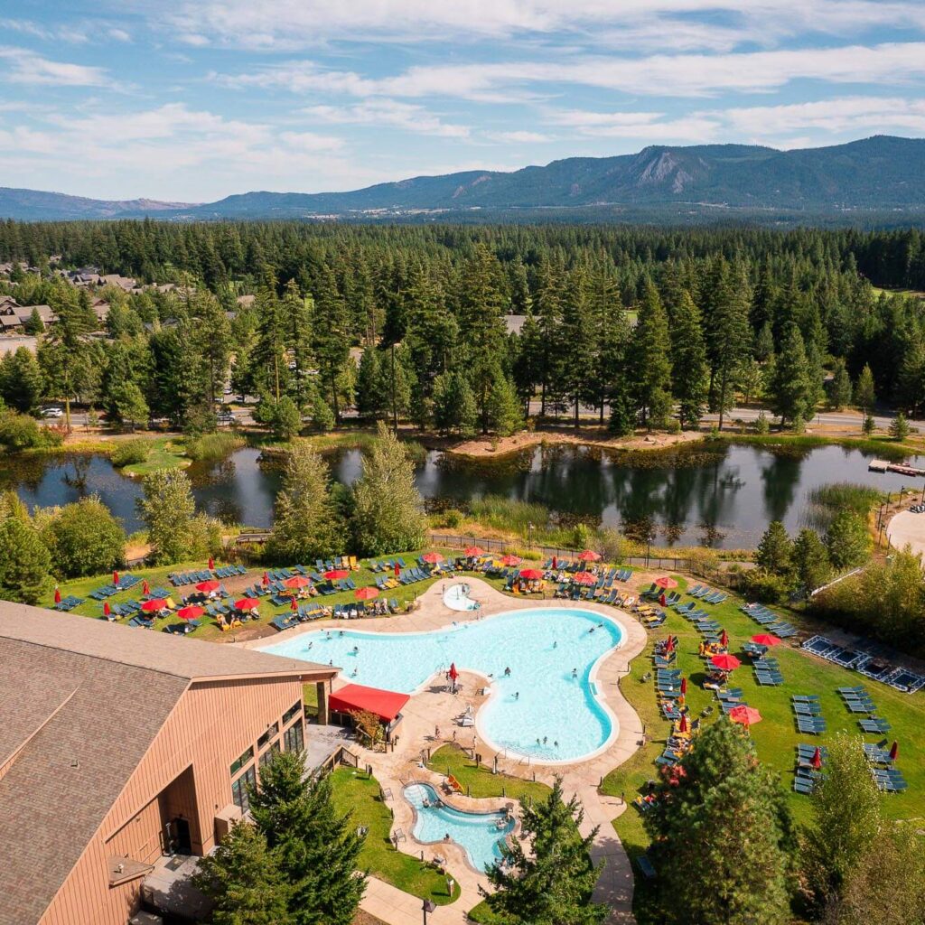 Aerial view of a resort pool surrounded by lounge chairs, umbrellas, trees, a pond, and mountains in the background on a sunny day.