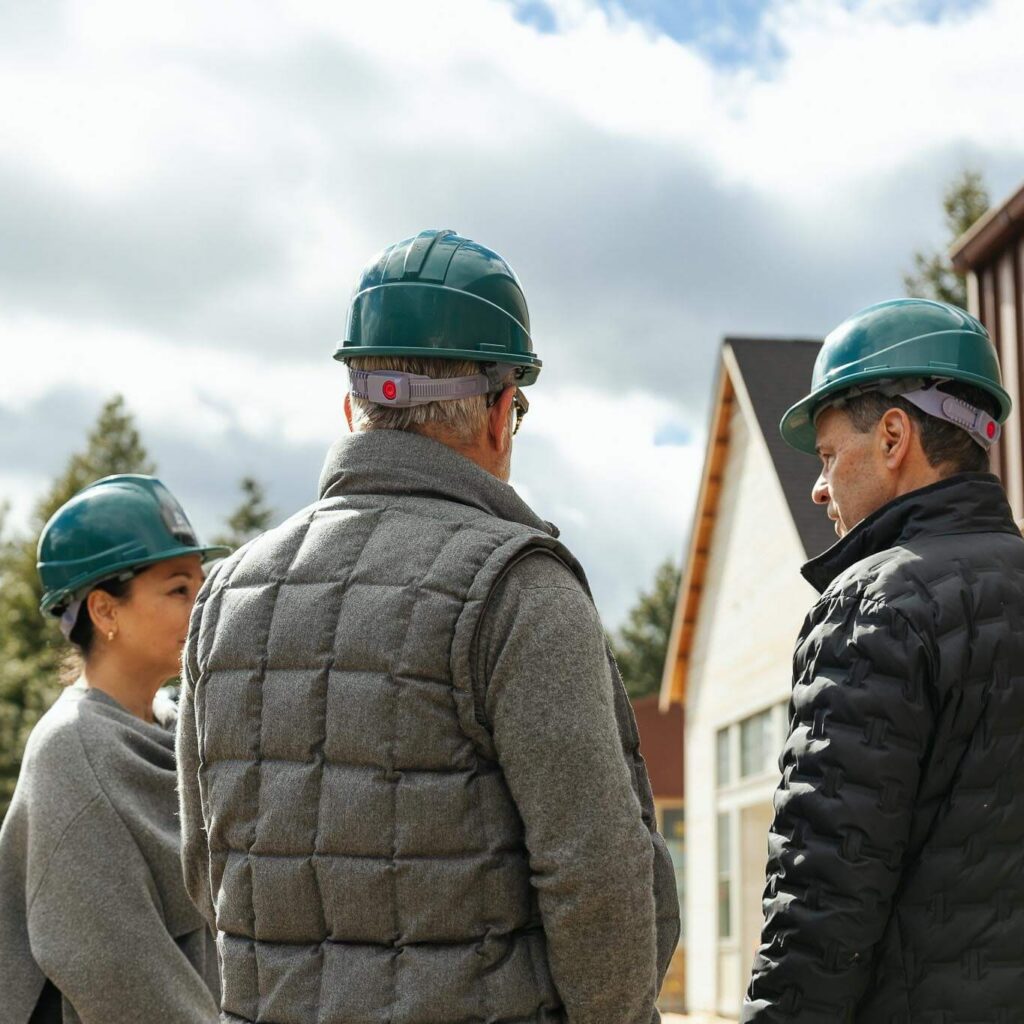 Three people wearing green hard hats and jackets stand outdoors at a construction site, facing away from the camera and talking. There are trees and a partially built house in the background under a cloudy sky.