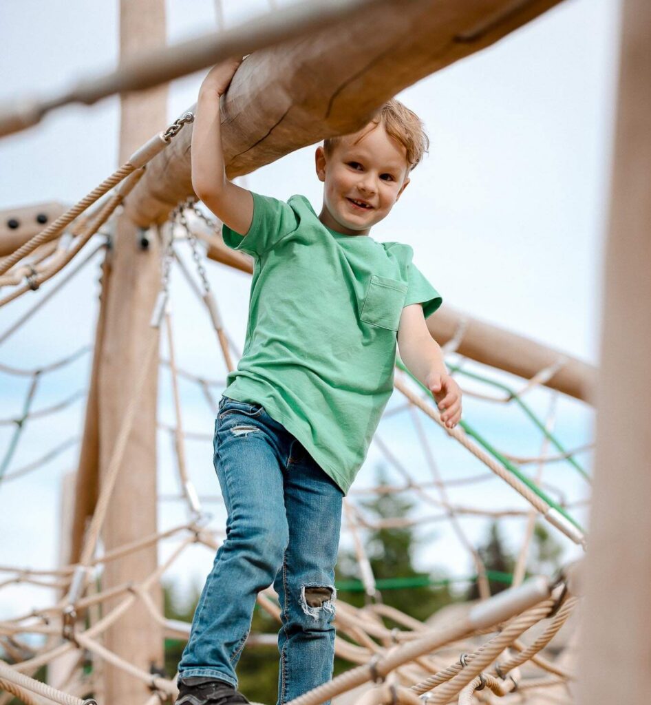 A young child in a green shirt and jeans smiles while balancing on a rope bridge at a playground, holding onto a wooden beam above for support.