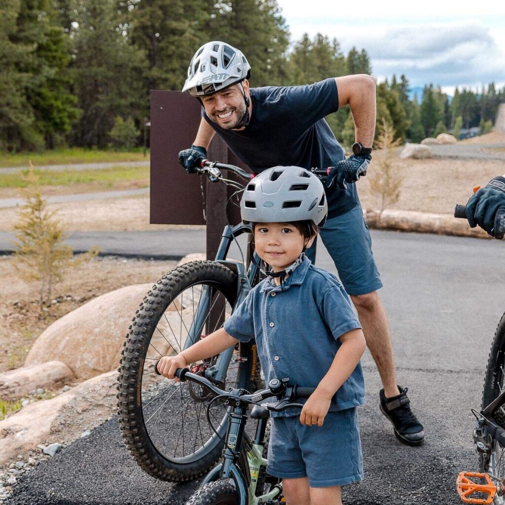 A man and a young boy wearing helmets pose with their mountain bikes on a path surrounded by trees. The boy stands with his bike while the man stands behind with his bike, both smiling.