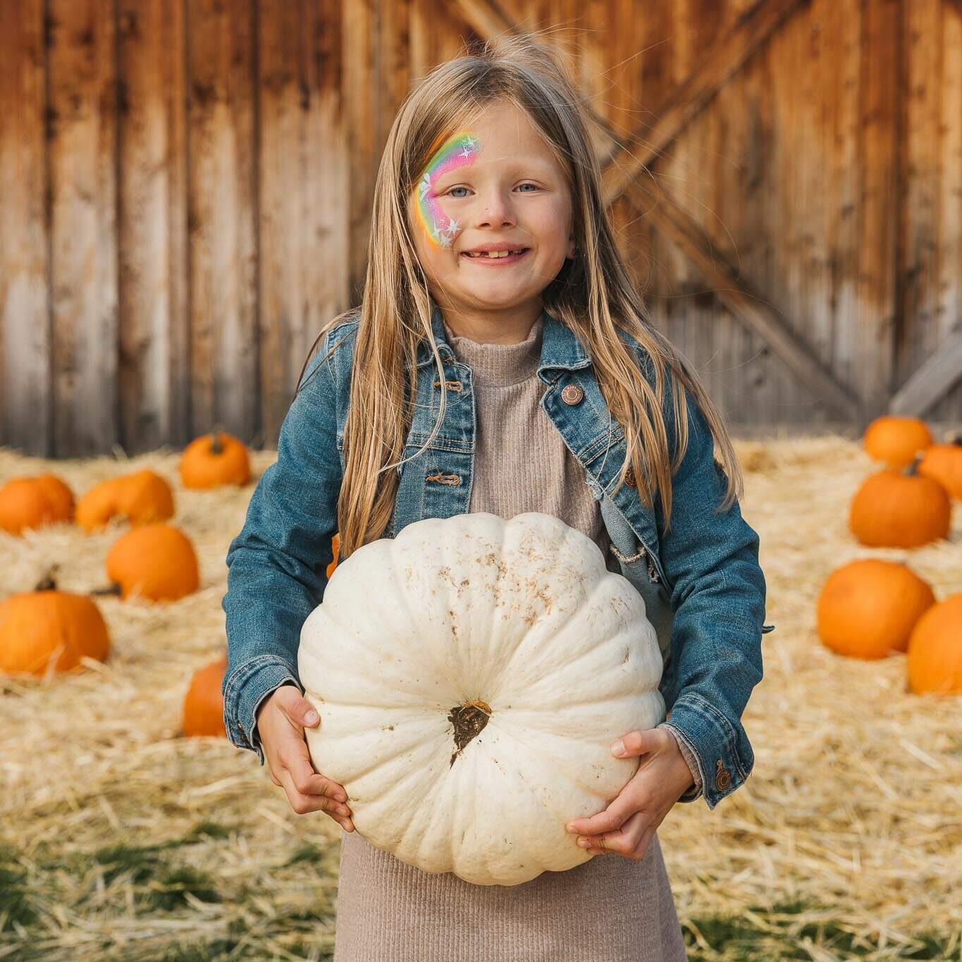 Smiling child with long blonde hair holds a large white pumpkin. She has colorful face paint and wears a denim jacket over a dress. Several orange pumpkins and a wooden barn are in the background.