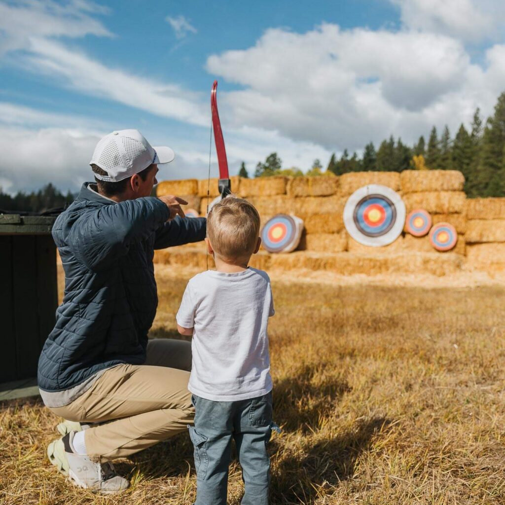 An adult kneels beside a child, helping aim a bow at several archery targets set up on hay bales outdoors on a sunny day.