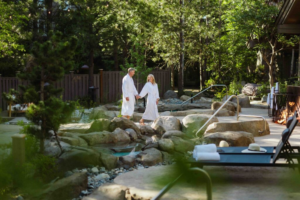 Two people in white robes walk hand in hand by an outdoor hot tub surrounded by rocks and trees, with lounge chairs and towels nearby in a tranquil, spa-like setting—an idyllic moment that captures the relaxing atmosphere of Spring events at Suncadia.