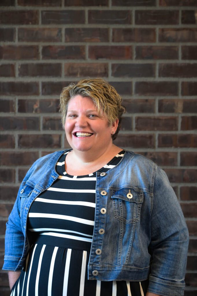A woman with short, blonde hair smiles while standing in front of a brick wall. She is wearing a black and white striped dress and a denim jacket.
