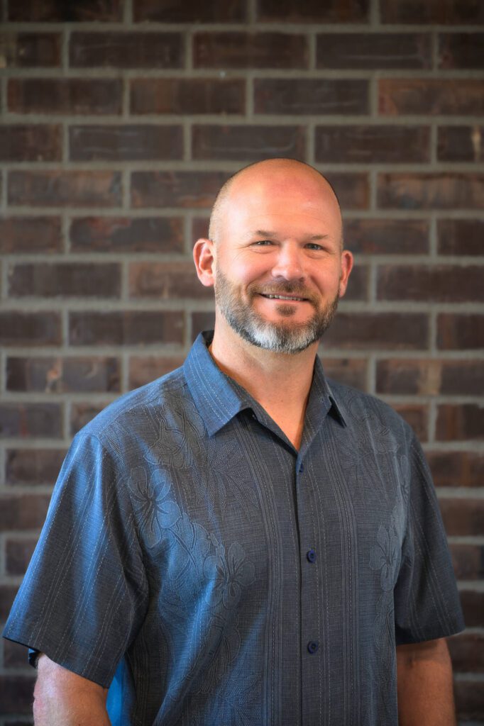 A smiling man with a bald head and trimmed beard wearing a short-sleeved, dark blue patterned shirt stands in front of a brick wall.