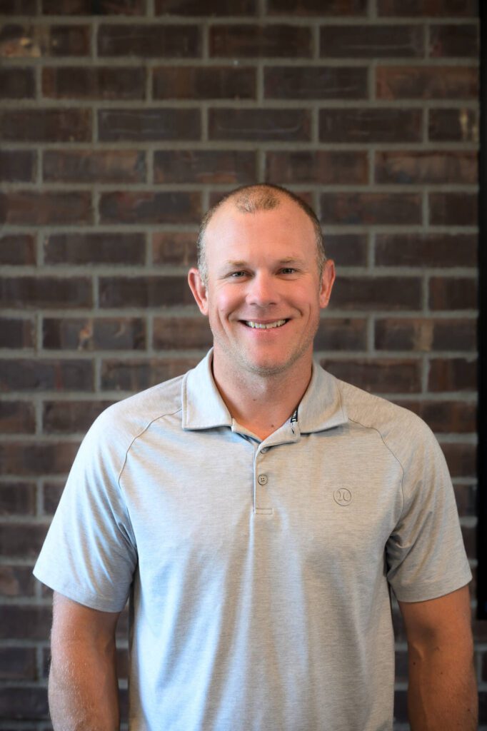 A man with short light hair, wearing a light gray polo shirt, stands smiling in front of a brown brick wall.