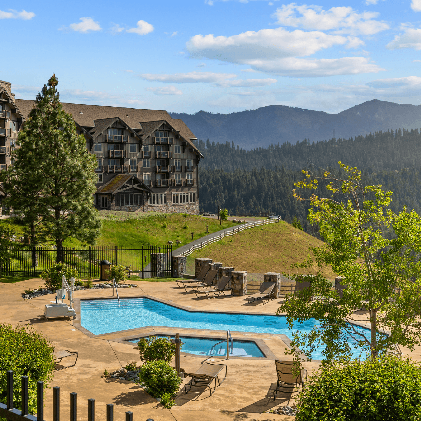 A scenic outdoor pool and hot tub at a resort overlook forested mountains under a blue sky, with lounge chairs, trees, and a large rustic building nearby.
