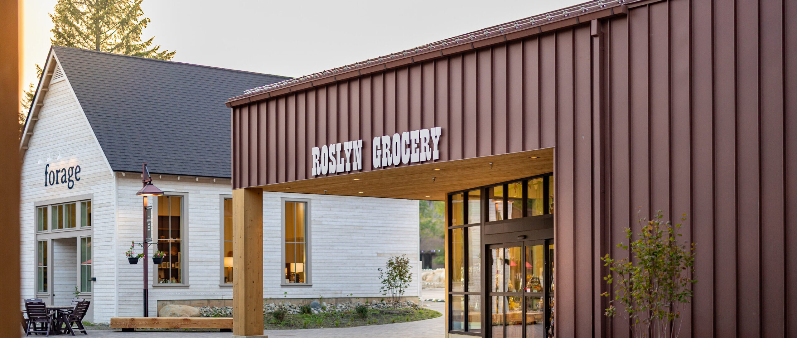 A modern grocery store with Roslyn Grocery on the sign sits next to a white building labeled forage, with benches, trees, and a paved walkway in front.