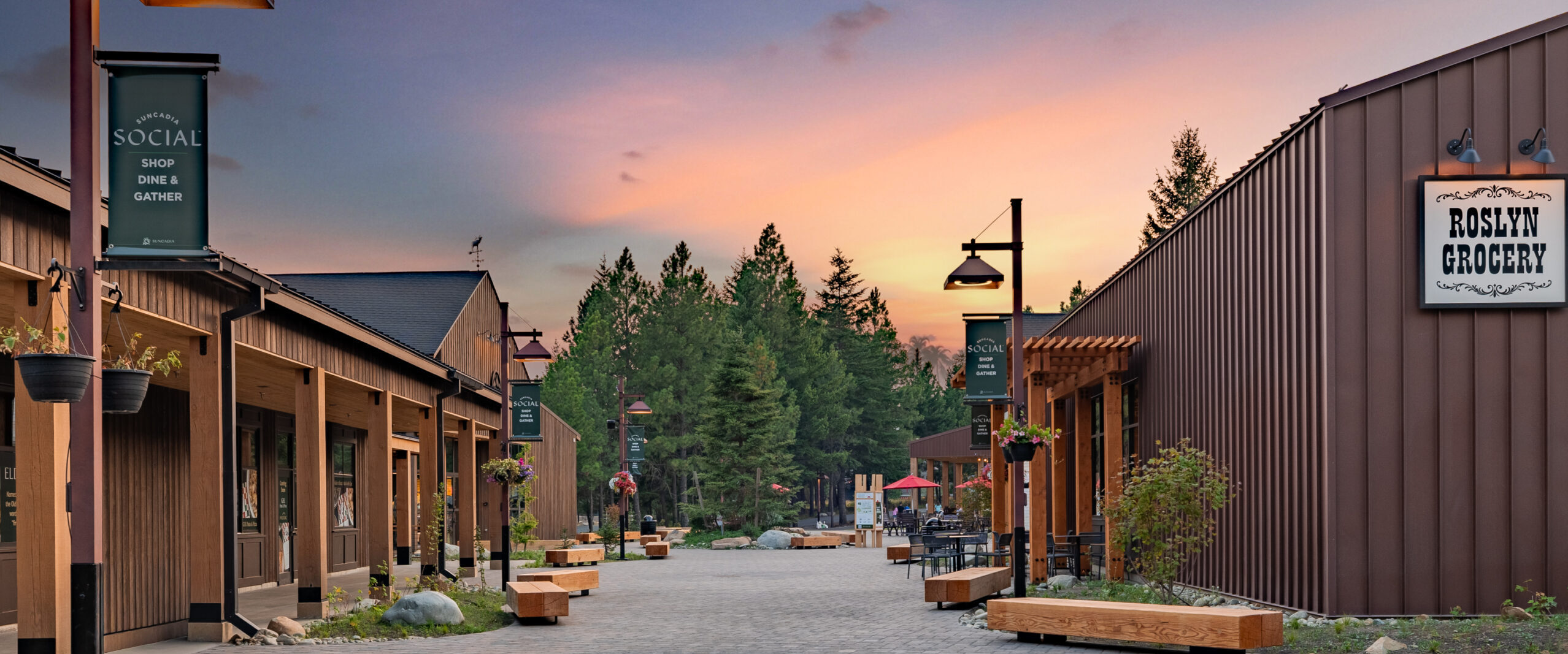 A quiet outdoor shopping area with wooden benches, trees, and shops, including Roslyn Grocery, at sunset. The sky is pink and orange, and no people are visible.