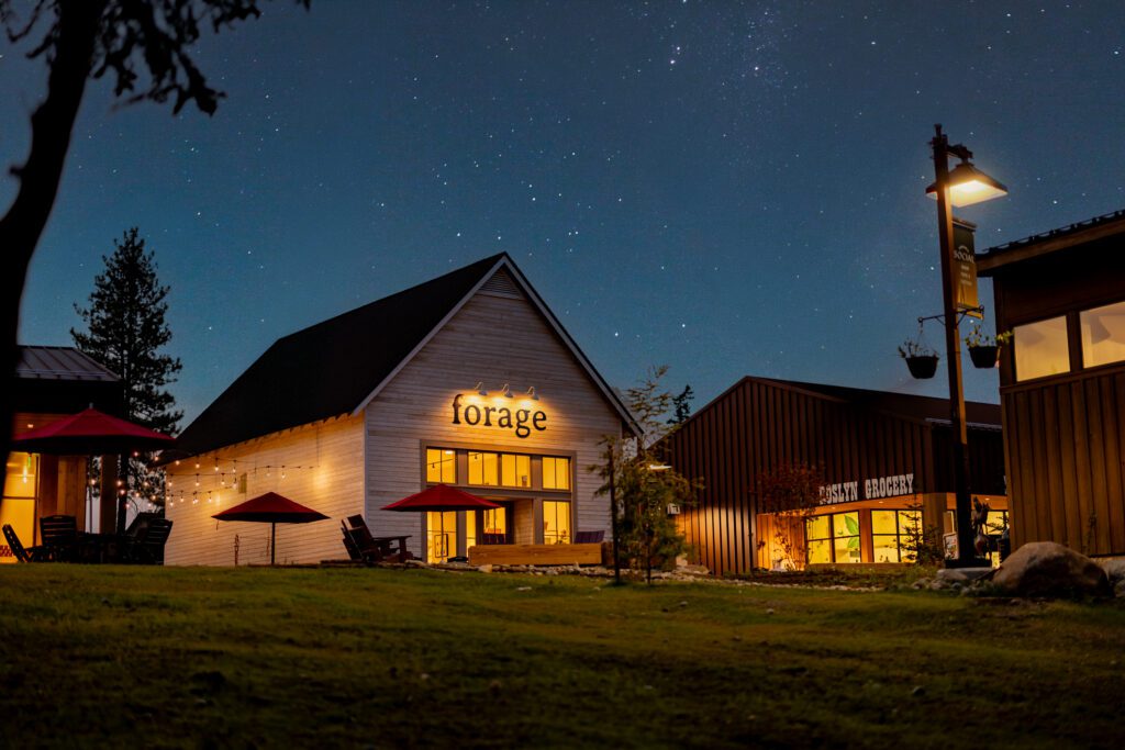 A warmly lit restaurant named forage sits under a starry night sky, surrounded by outdoor seating, string lights, and nearby buildings on a grassy lawn.