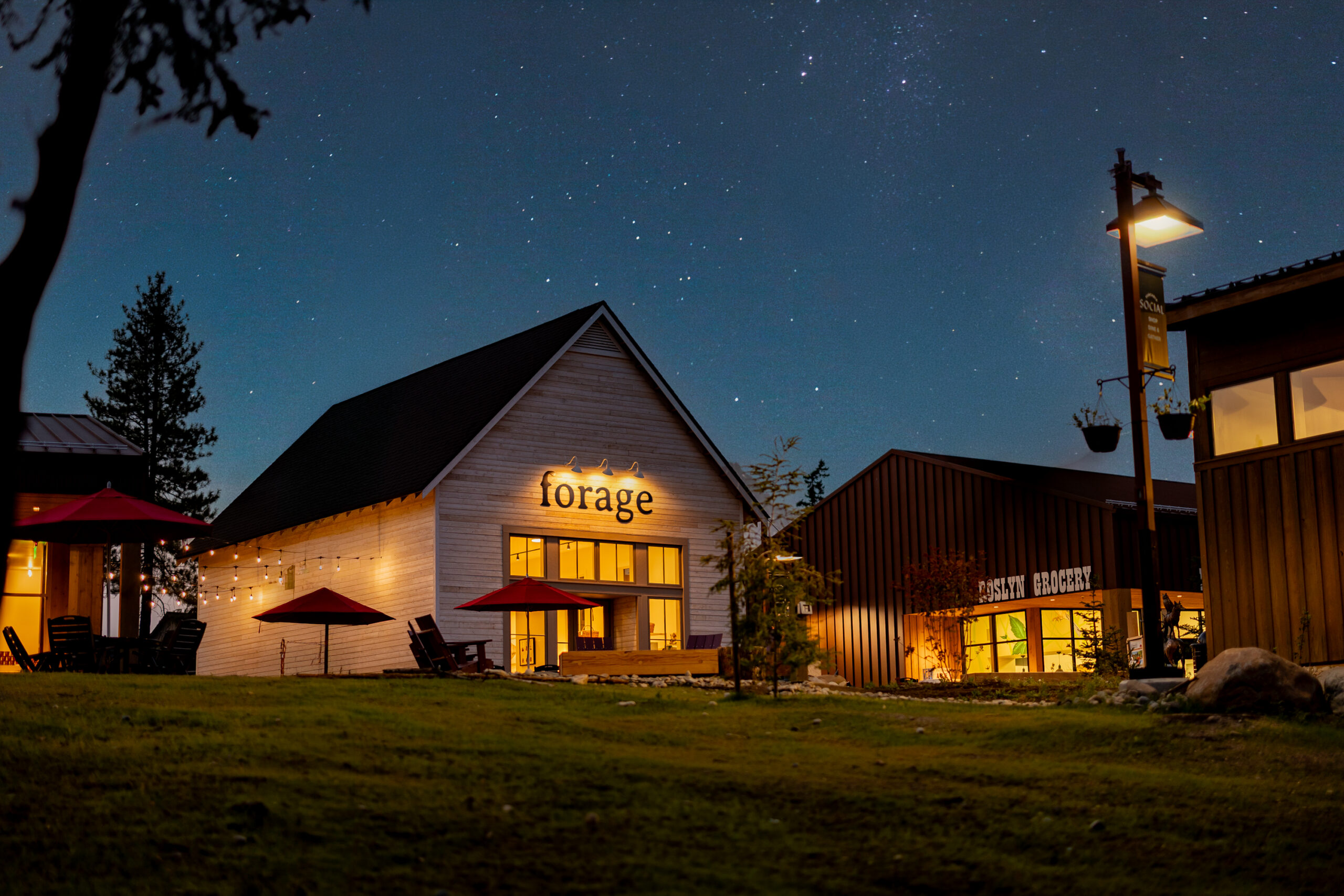 A warmly lit restaurant named forage sits under a starry night sky, surrounded by outdoor seating, string lights, and nearby buildings on a grassy lawn.