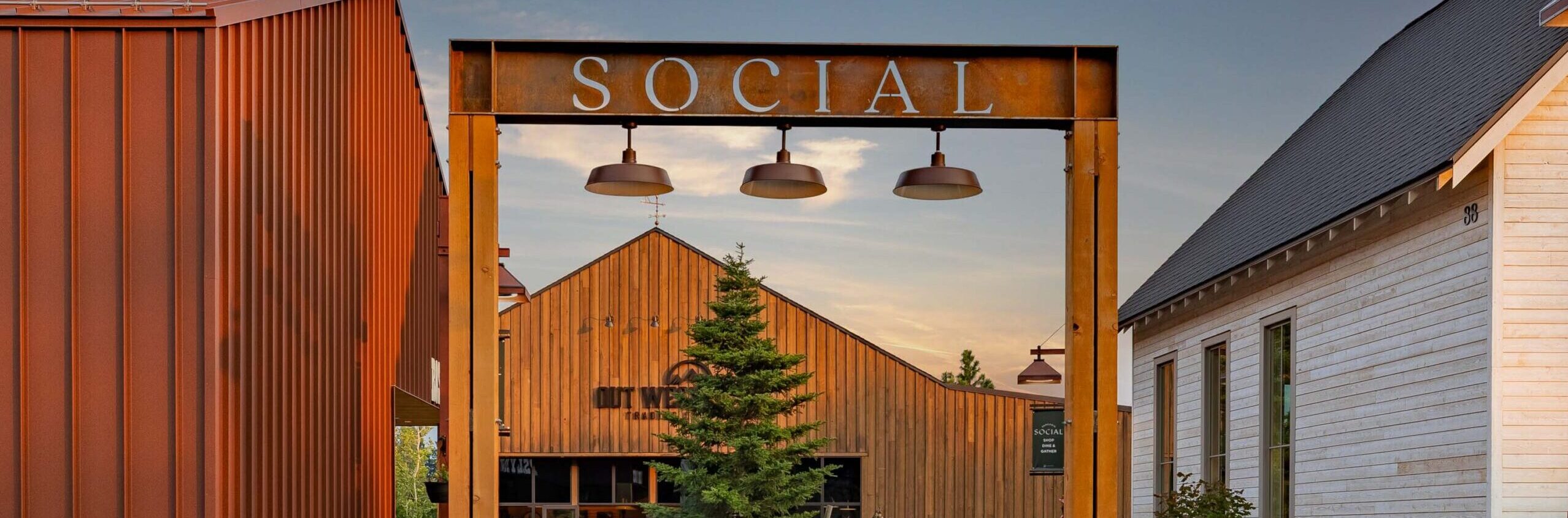 A metal archway labeled SOCIAL stands between modern barn-style buildings, with a pine tree and people visible through the entrance on a paved path at sunset.